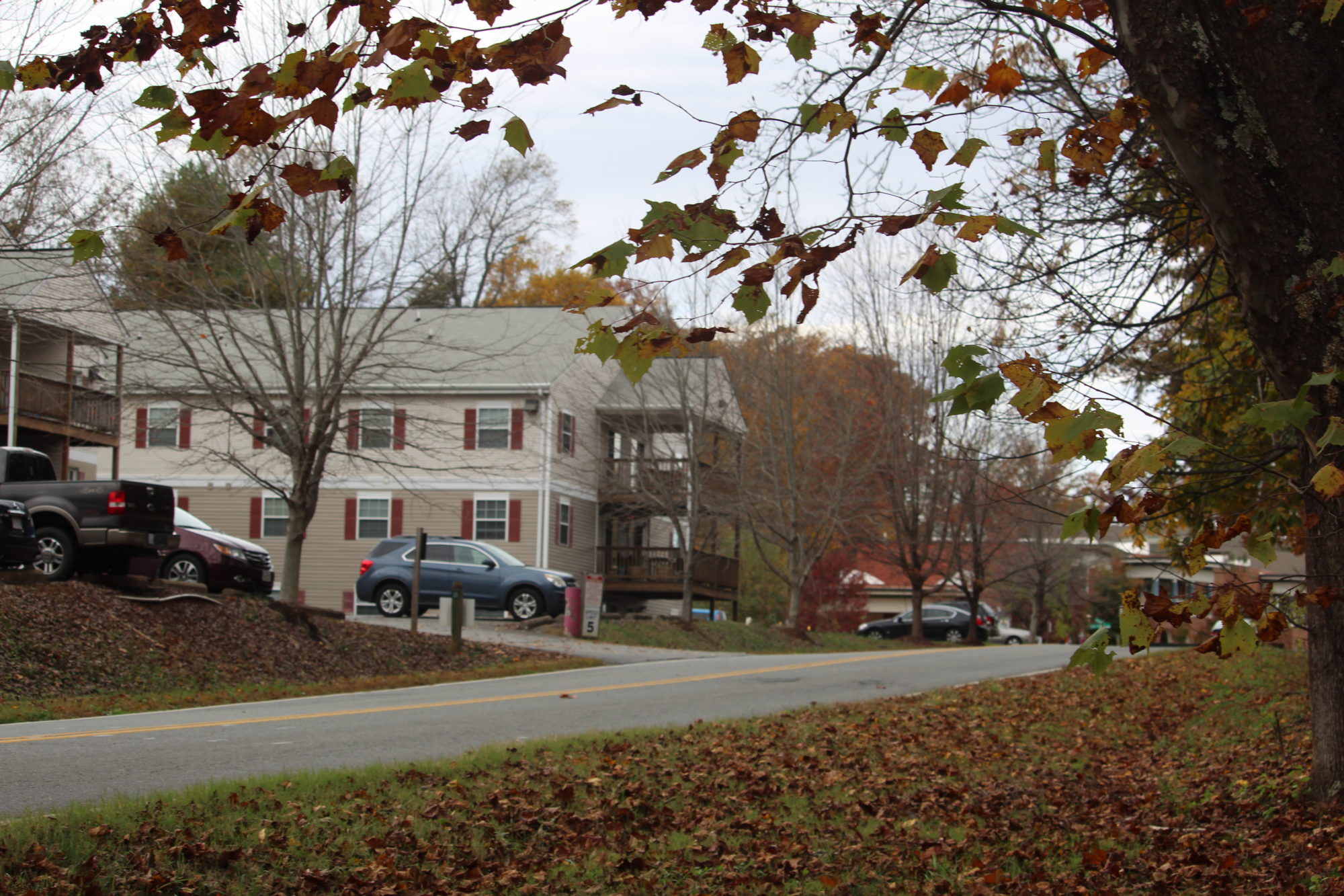 A three-story building seen from a grassy area across a two-lane road. The building has beige panels and windows with shutters, and wooden balconies. Cars are parked in a lot next to the building.