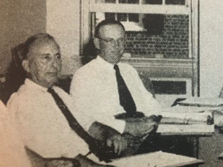 A black and white photo of two mean in white shirts and dark ties sitting at a table.