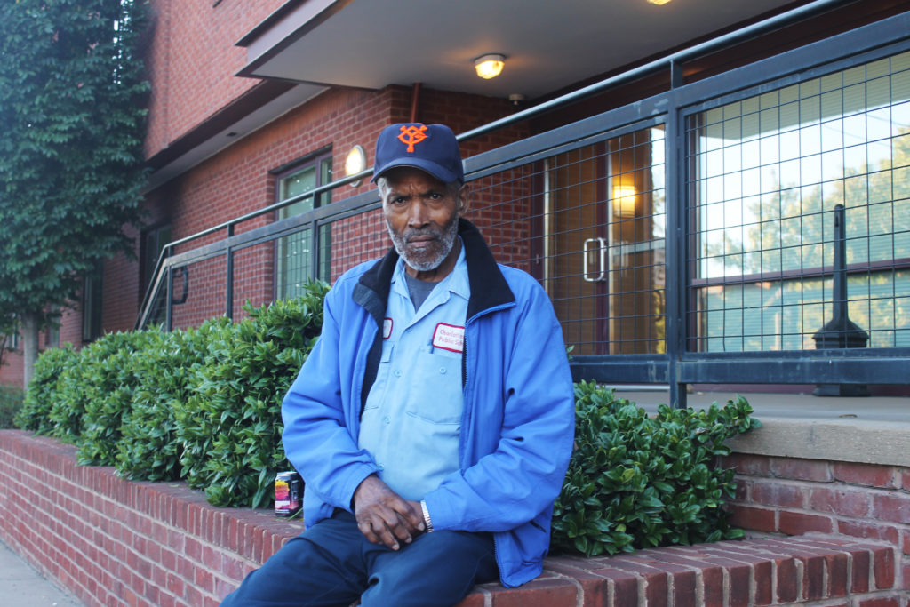 A man in a blue jacket and baseball cap sits on the edge of a brick planter in front of a building.