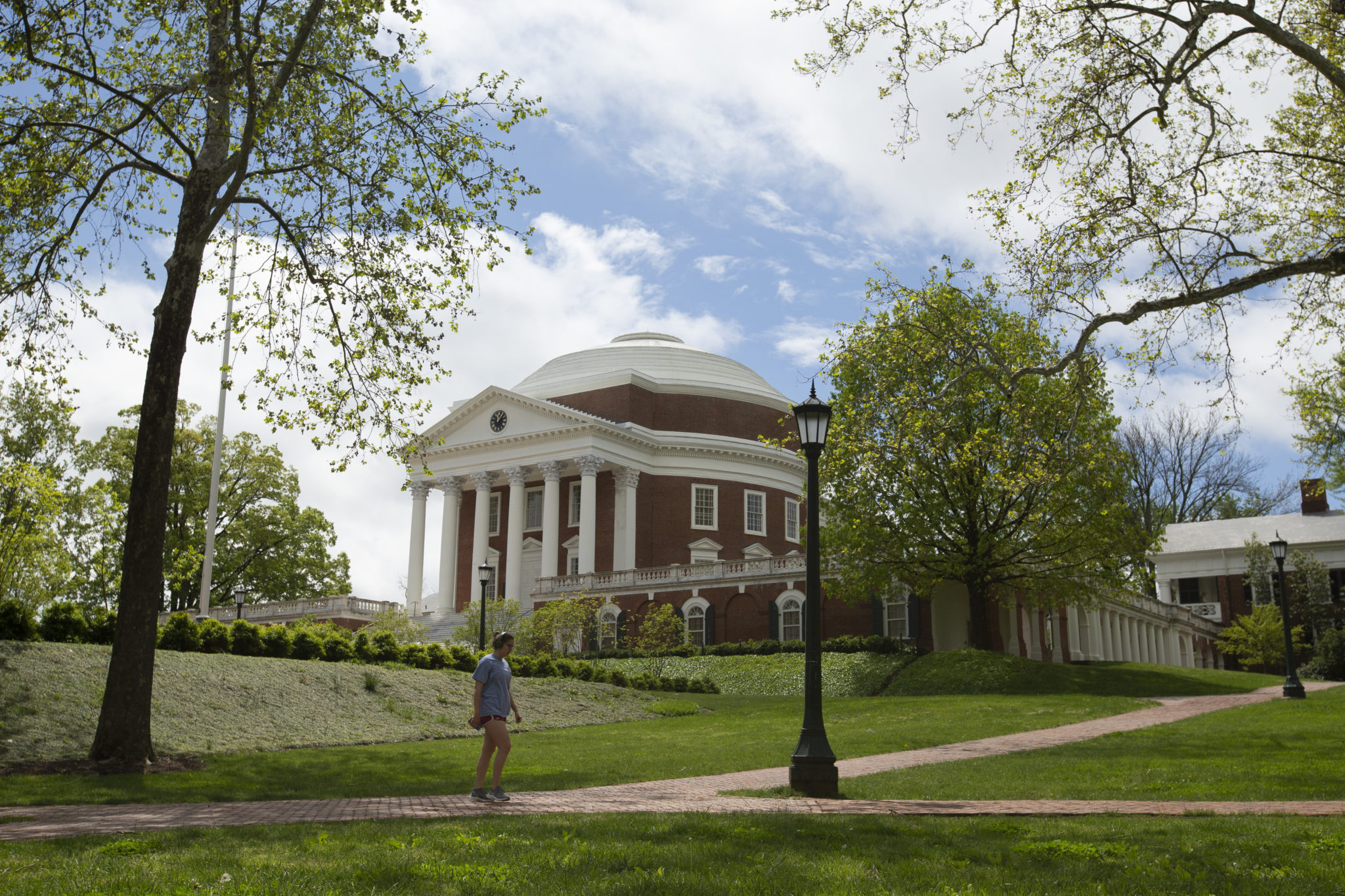 A person wearing headphones walks in front of a brick building with columns and a round roof, a green lawn and trees around.