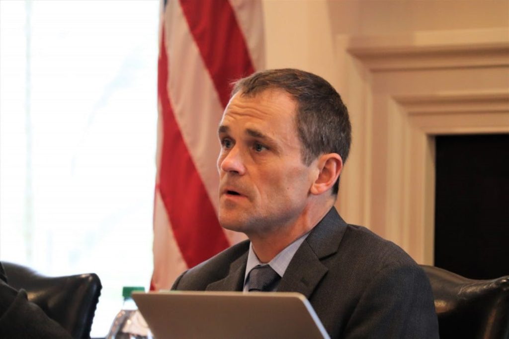 A man in a suit sits in front of an American flag and a white fireplace.