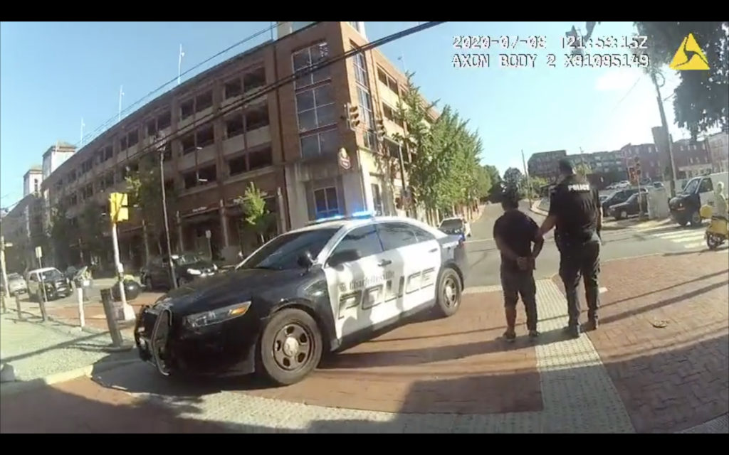 Footage from a body worn camera shows a police officer holding onto a man who has his hands cuffed behind his back as they approach a vehicle.