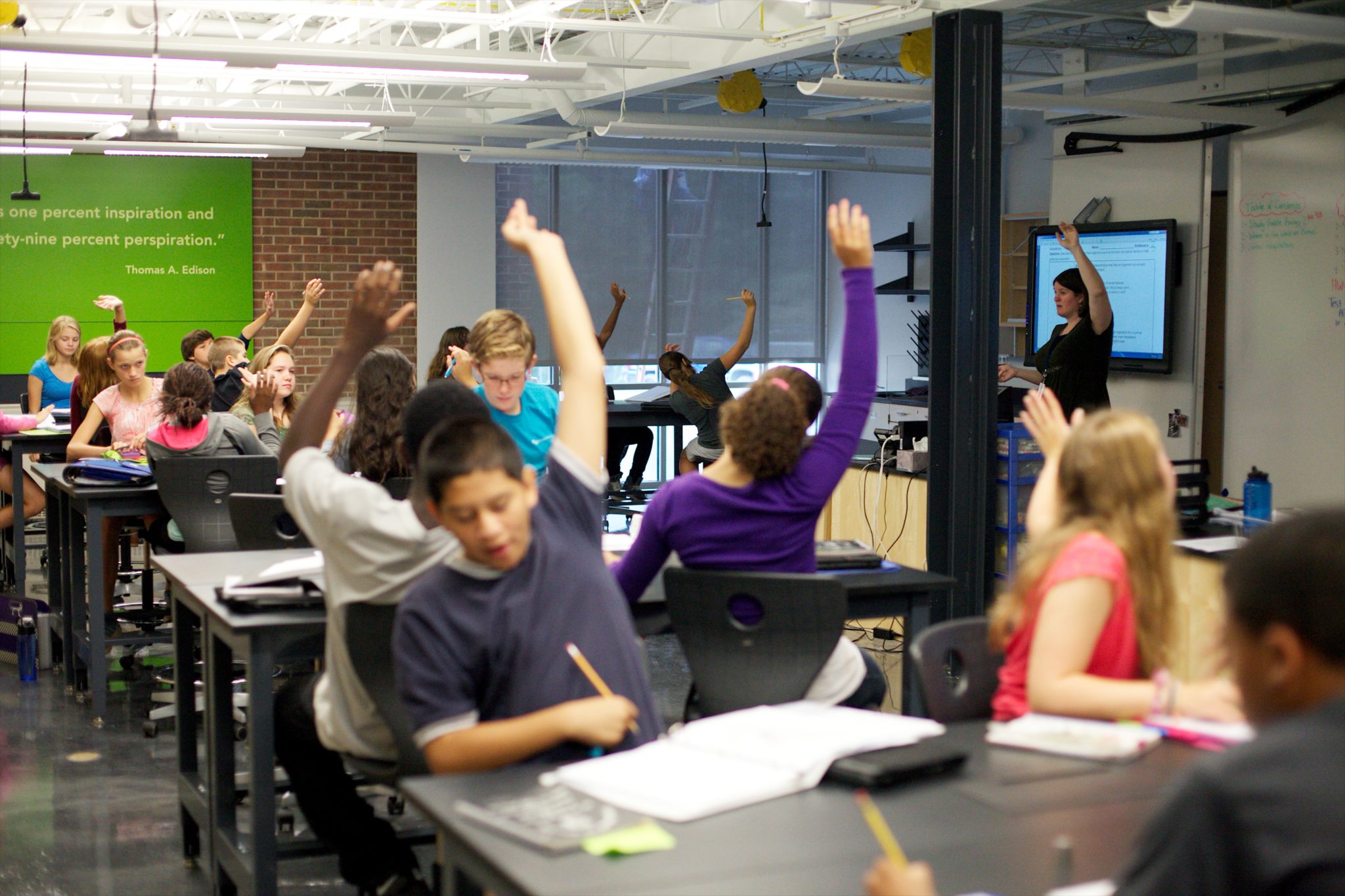 classroom of students at grey tables raise their hands, with a teacher off to the side in front of a screen raising her hand