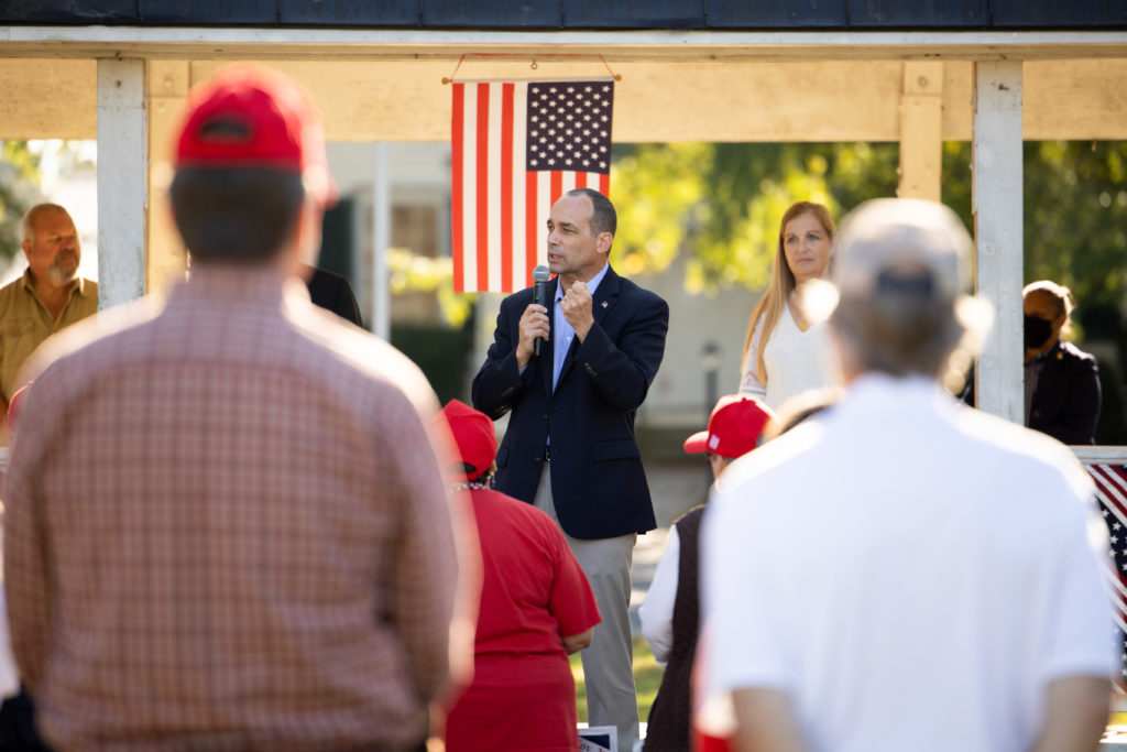 A man in a suit jacket speaks outside into a microphone. Behind him is an American flag. In front of him are two men watching.