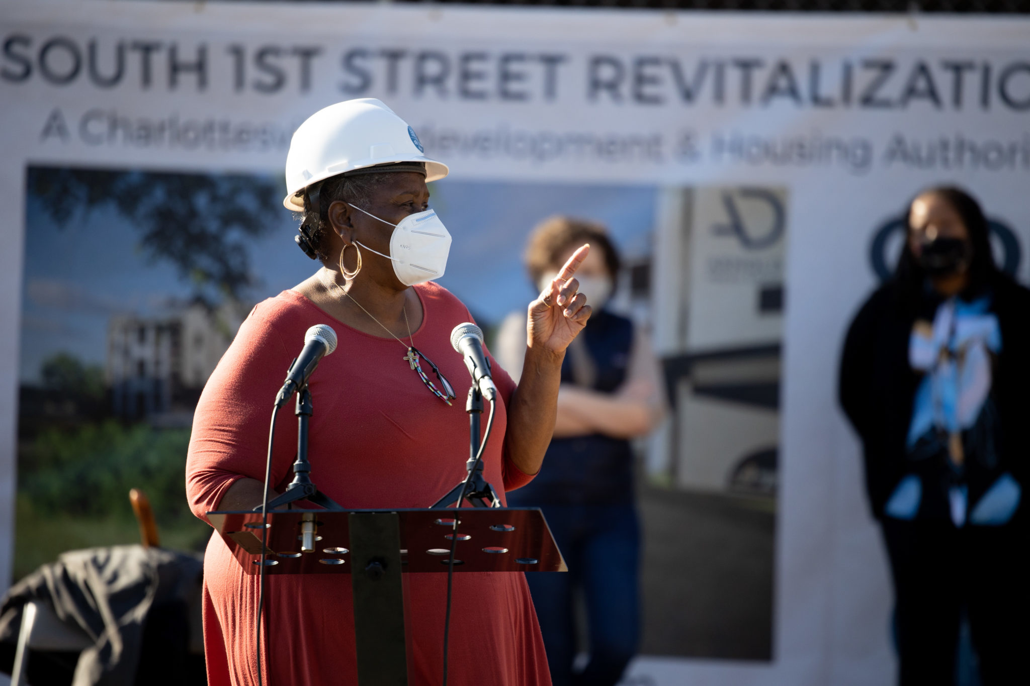 Woman in hard hat and mask speaks into microphone with sign behind her that reads, "South Street Revitalization"