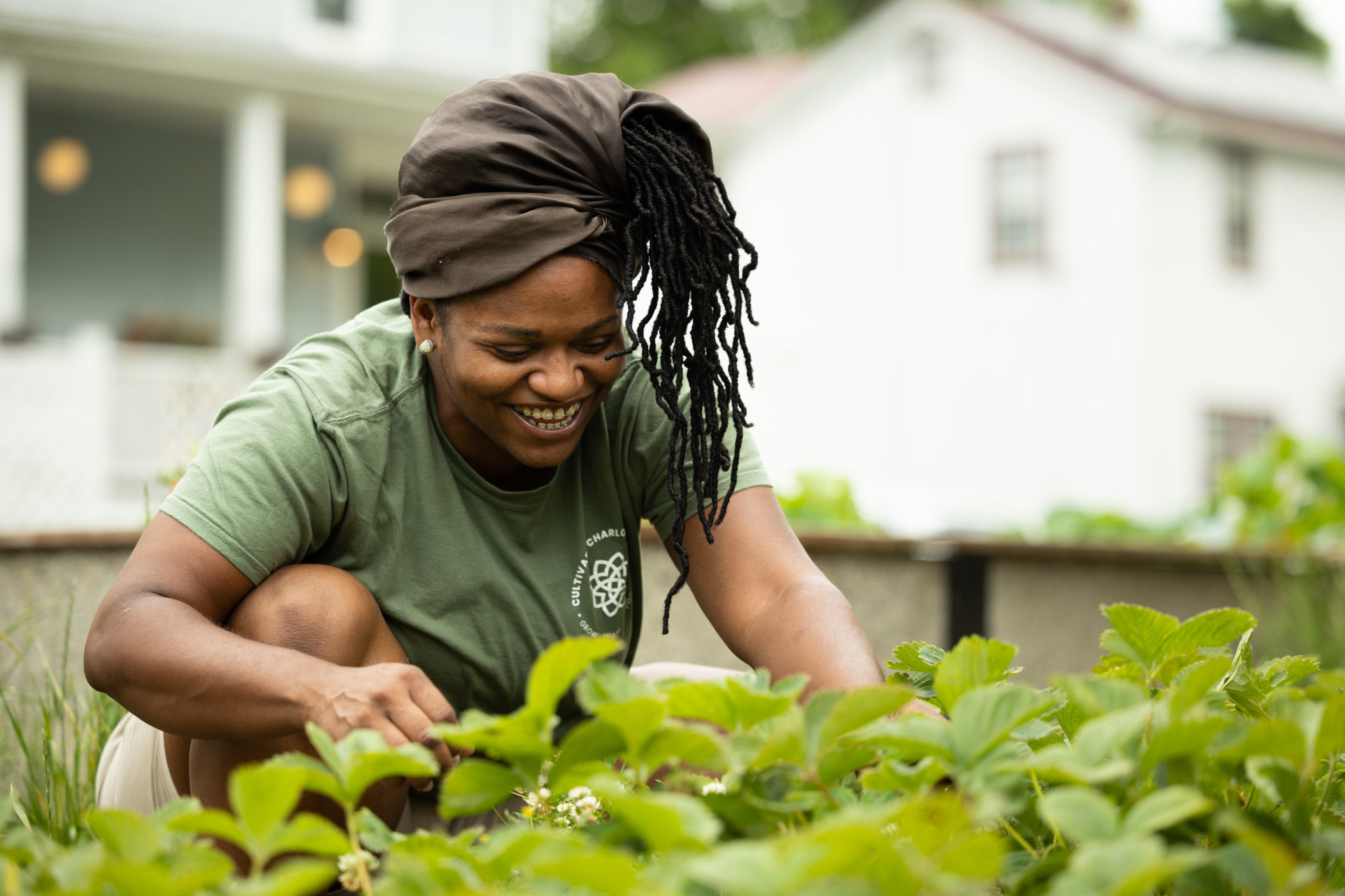 A woman kneeling in a strawberry patch. She's smiling as she pulls weeds and looks for fruit to pick.