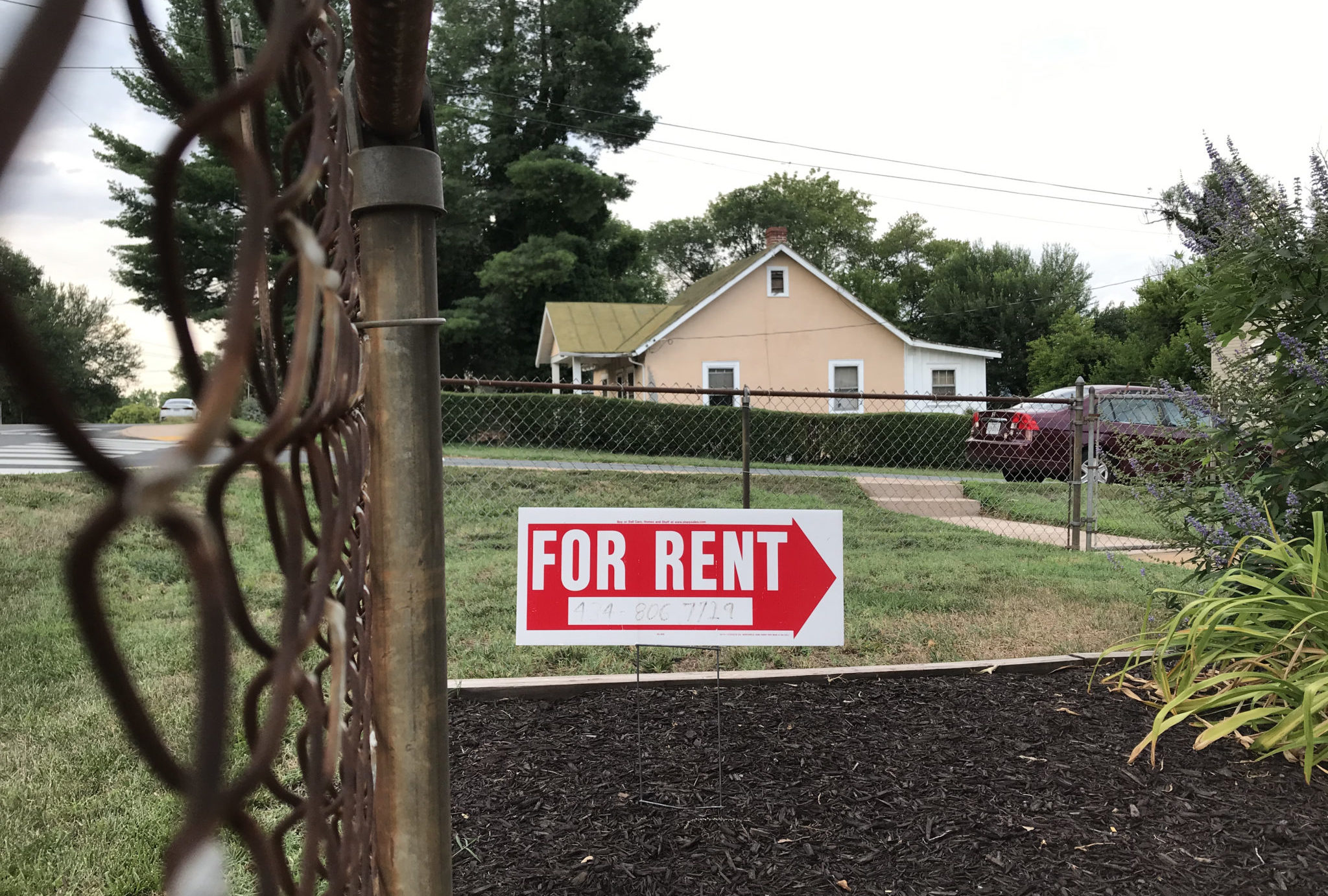 A sign reading "For Rent" in all capital letters is stuck into mulch in a front yard, behind a chain link fence. In the background, a car and a house.