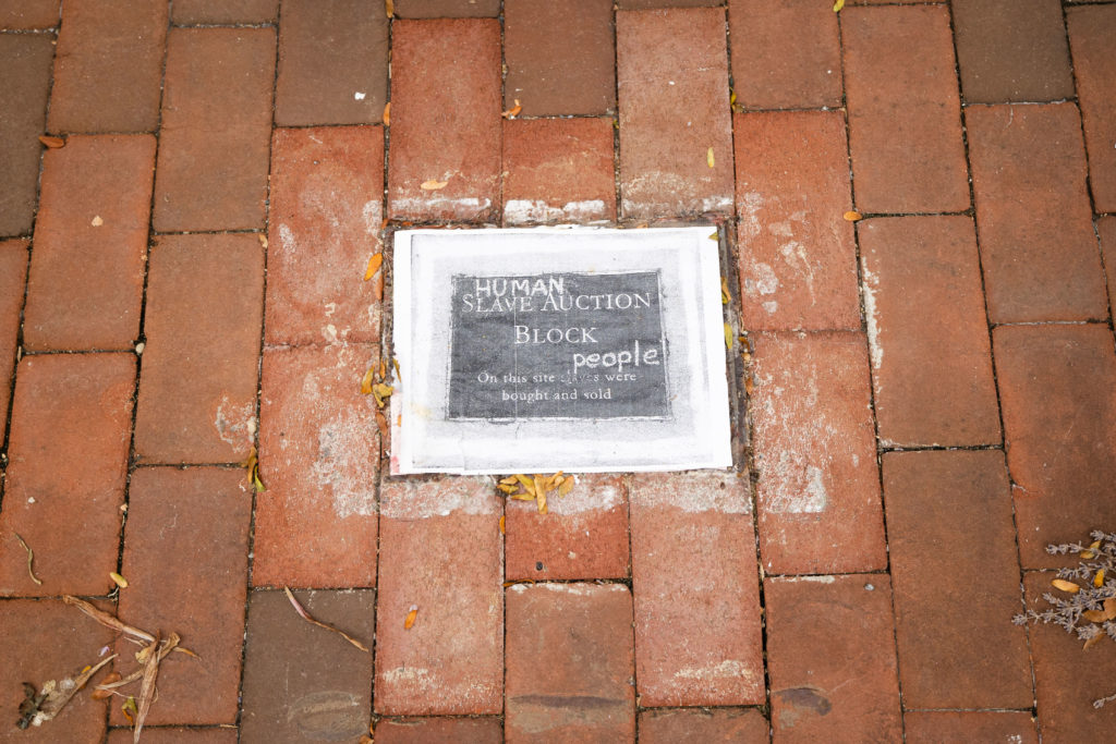 A photograph of a brick sidewalk. In the middle of the photo, embedded in the sidewalk, is a piece of paper with a black and white image of a plaque on it. The plaque reads, "Slave Auction Block: On this site slaves were bought and sold." Someone has added words to it, intending to replace "Slave" with "Human" and "slaves" with people.
