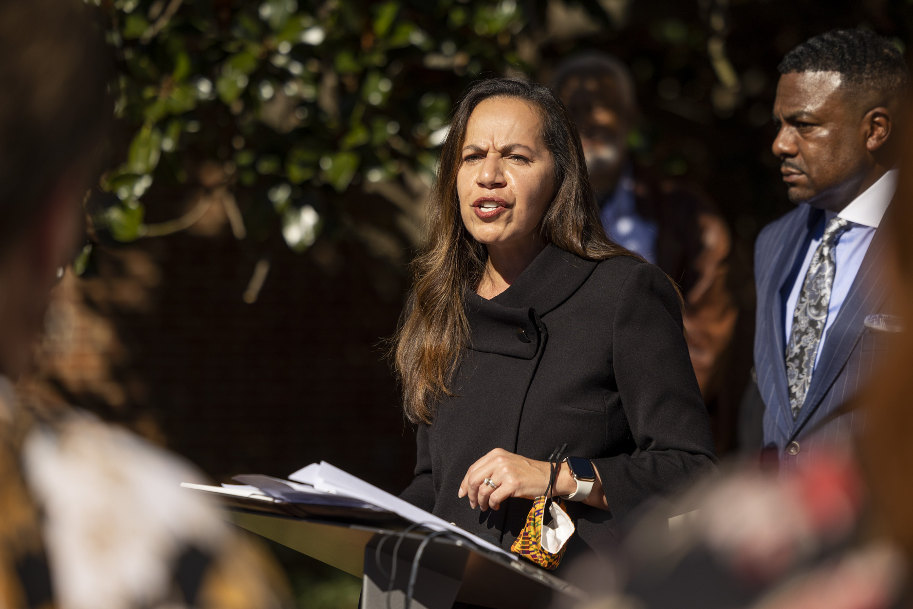 A woman stands outside at a podium speaking. A man in a suit stands behind her.