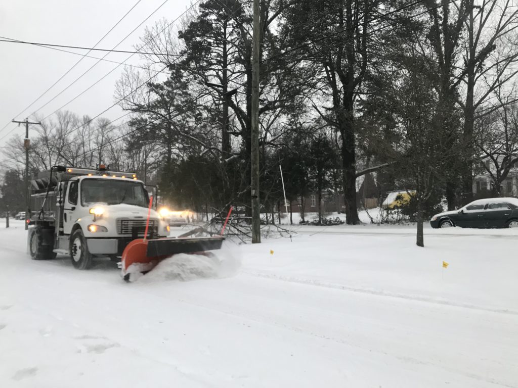 A snow plow with its headlights on moves about six inches of white snow off a major road at dusk.