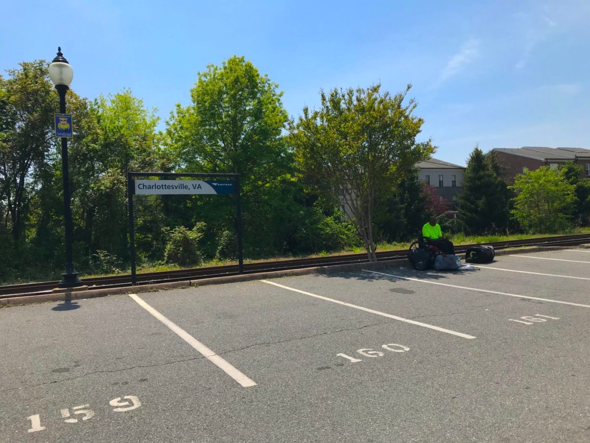 A person in a wheelchair sits alone in the shade of a small tree in a paved parking lot. A nearby rectangular sign reads “Charlottesville, VA Amtrak.” There are train tracks next to the parking spaces, and some trees and apartment buildings in the background.