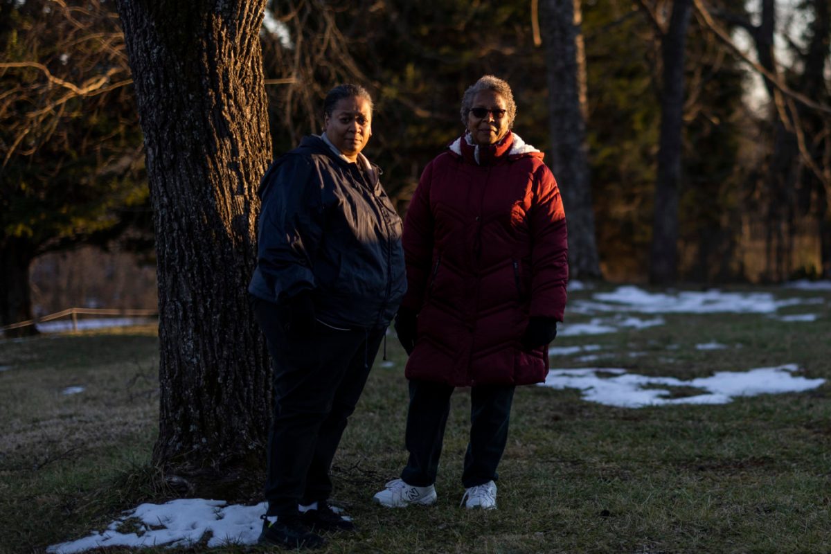 Two women stand near a tree in a grassy area.
