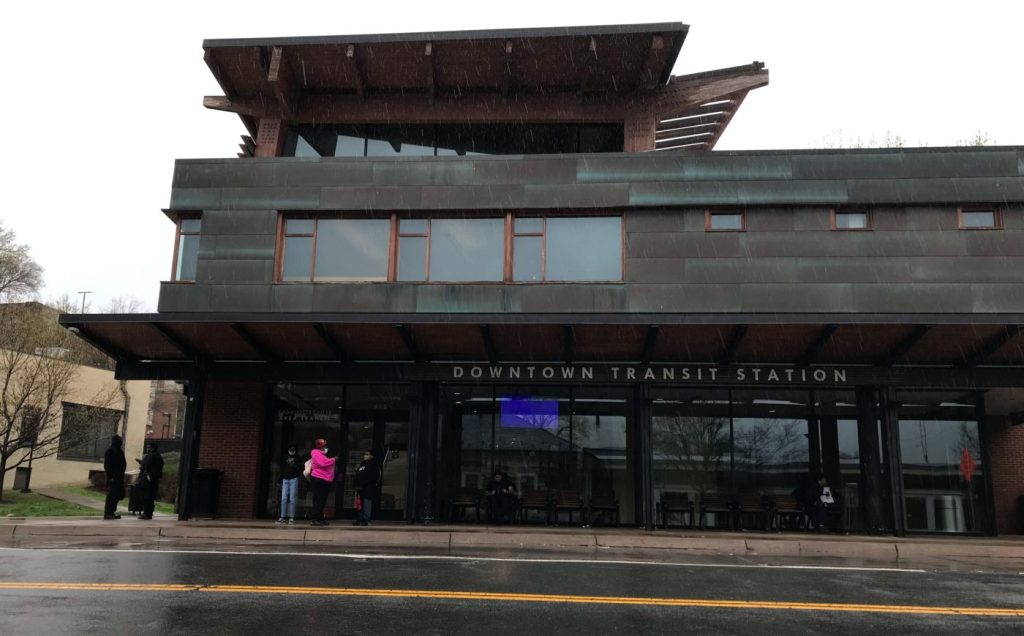 Several people wait in front of a two-story building on a rainy day. Building sign reads: “DOWNTOWN TRANSIT STATION”