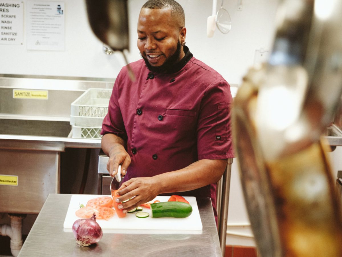 A man in a red chef’s shirt chops vegetables at a stainless steel table. In the foreground stainless steel frying pans hang from the ceiling.