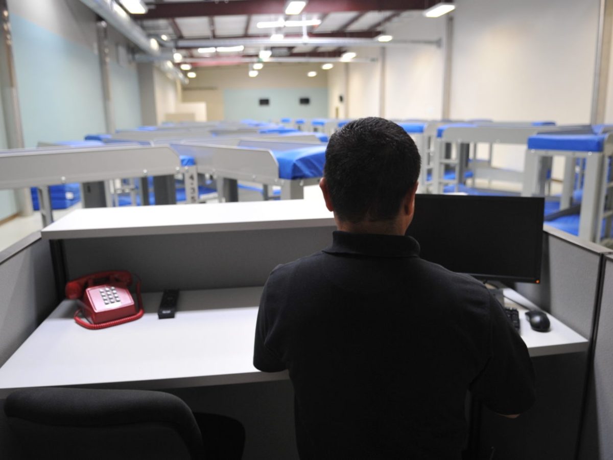 A man stands at a workstation, his back to the camera, with a monitor and red phone on a gray desk. Beyond there are many bunk beds in star patterns with blue mattresses.