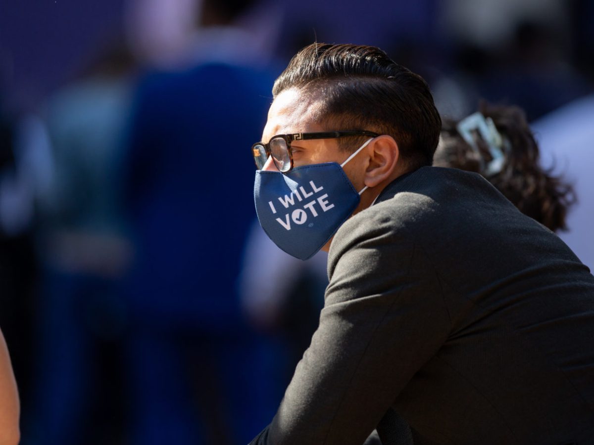 A man with glasses and a black blazer looks off camera. He is wearing a blue face mask that reads “I will vote.” There are other people whose faces aren’t shown around him.