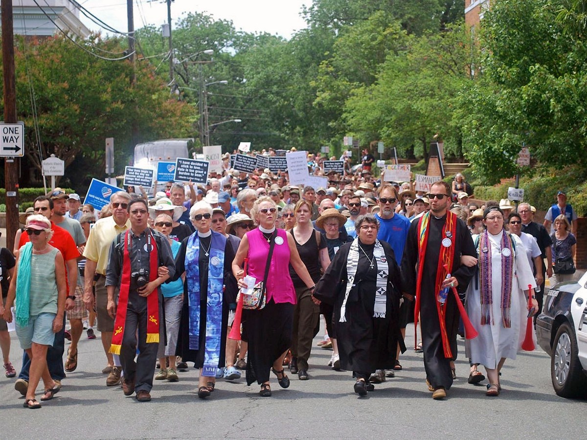 Religious leaders march in front of a crowd down a street, with trees behind them and a police vehicle in the corner of the image.