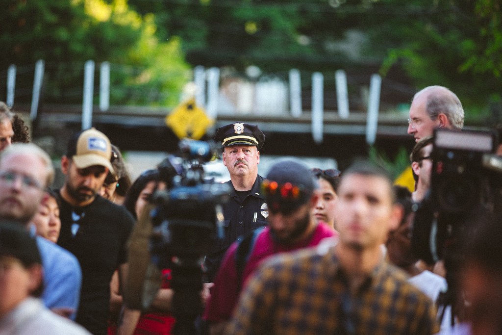 At the center of the photo is a police officer wearing a dark uniform and a hat. He is surrounded by people, some of them with large cameras, and all of their faces are blurred.
