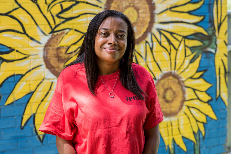 A woman in a pink shirt poses for a picture in front of a mural with yellow flowers painted onto a blue background.