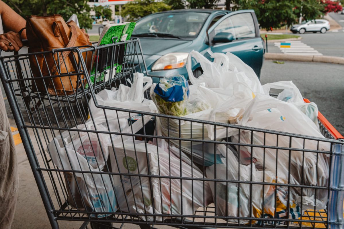 A close-up image of a large shopping cart full of plastic bags containing salad mix, celery, a loaf of bread, boxes of crackers, and other items. The stack of bags is high enough to peek over the edge of the cart.