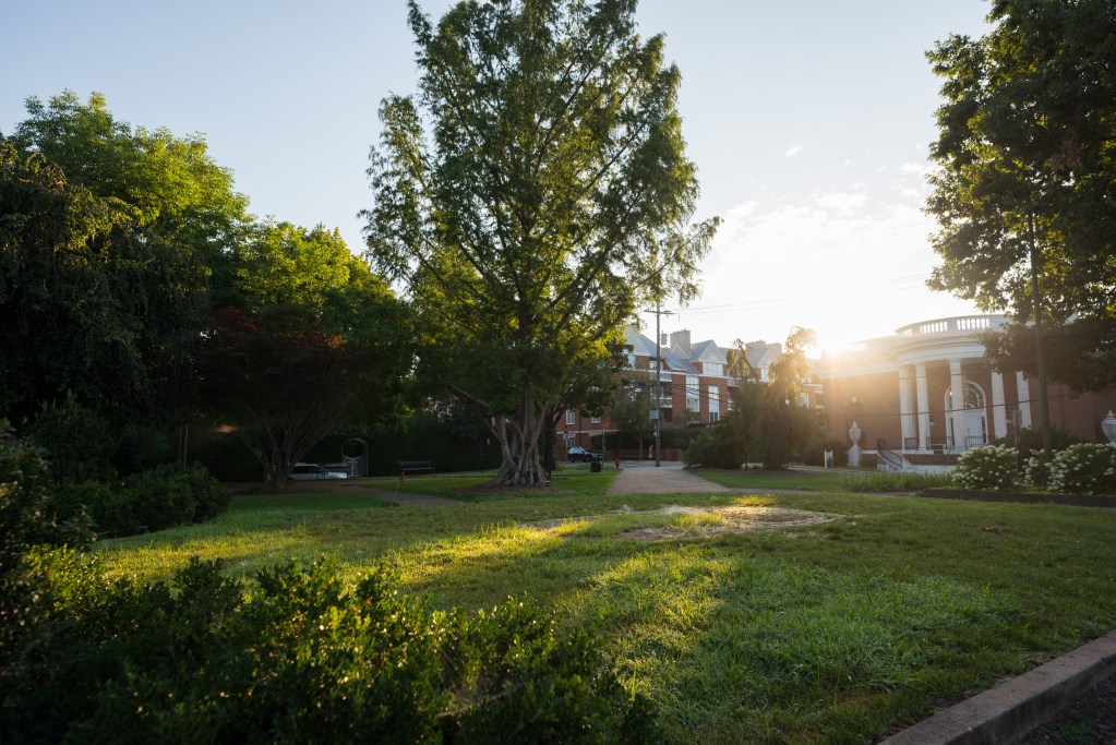 A green space with grass and trees is shown at sunrise. In the background are red and white brick buildings.