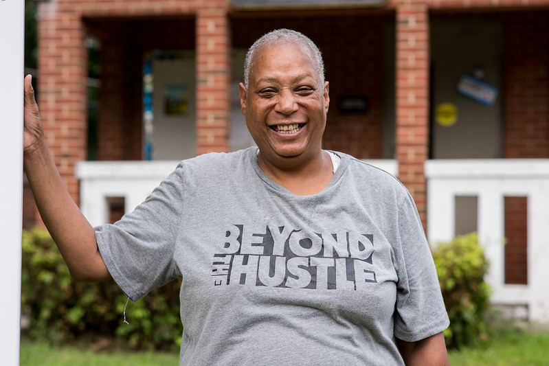 A woman in a gray t-shirt smiles for the camera in front of a brick building with columns.