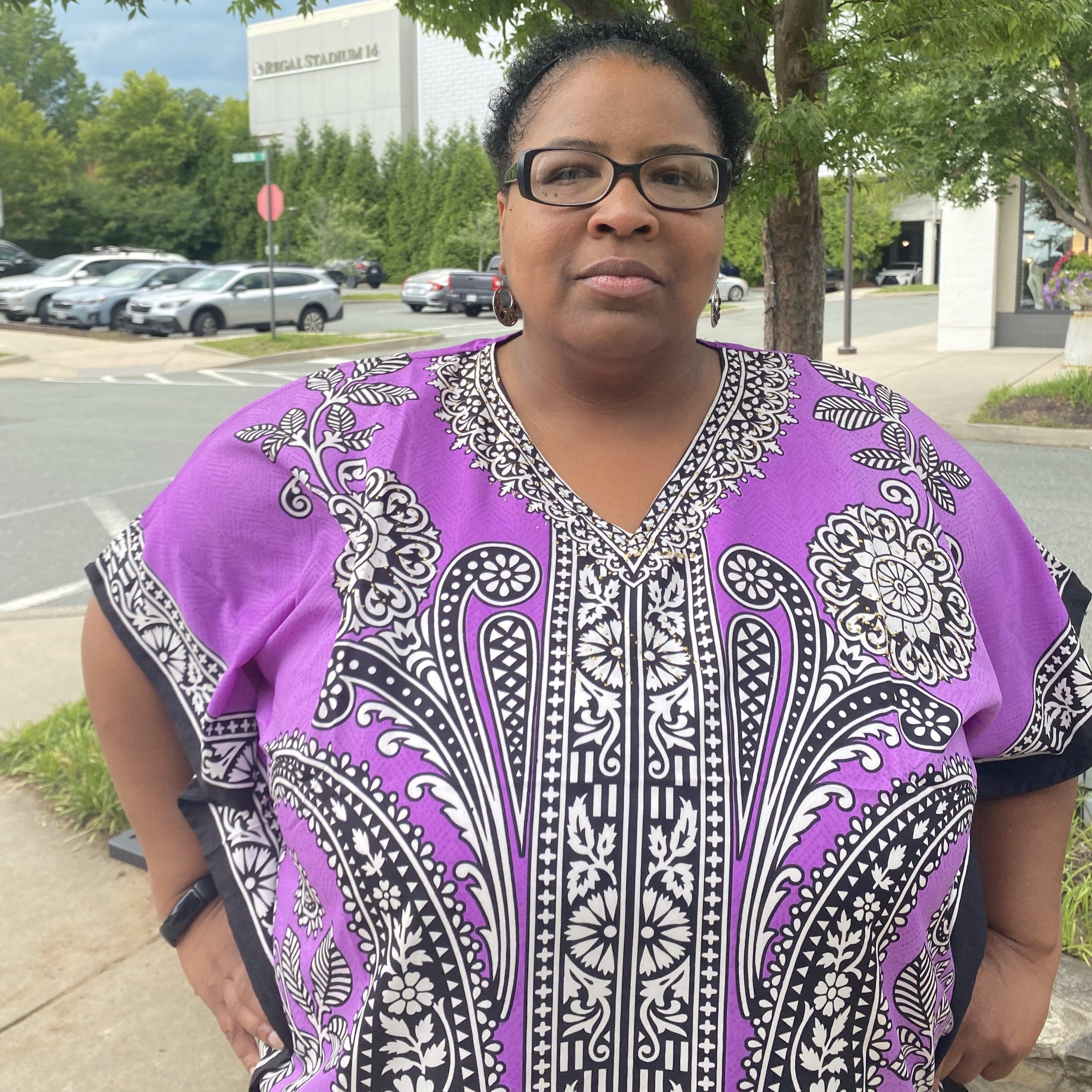 A woman in a pink shirt and glasses poses for a picture in a parking lot in front of a small tree.