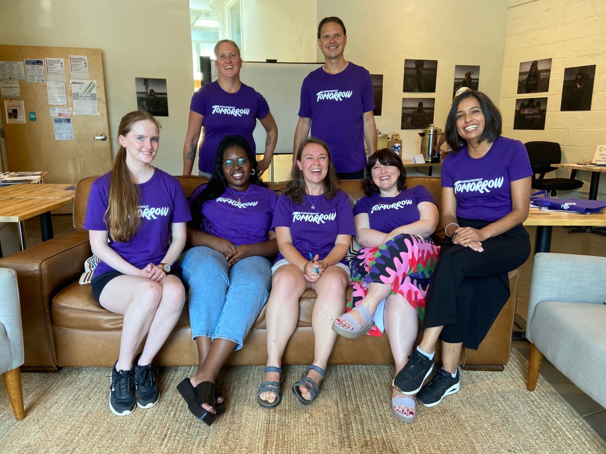 A group of seven people in purple t-shirts, with the logo of Charlottesville Tomorrow, pose on and behind a brown sofa.