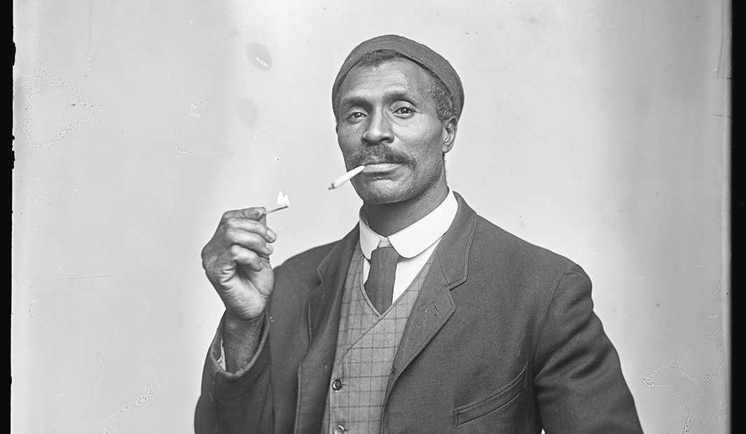 A black and white photograph shows a man sitting down and looking directly at the camera. He wears slacks and a jacket, a plaid waistcoat, a white shirt and tie. His left hand sits in his lap and his right hand holds a match to a cigarette dangling from his mouth.