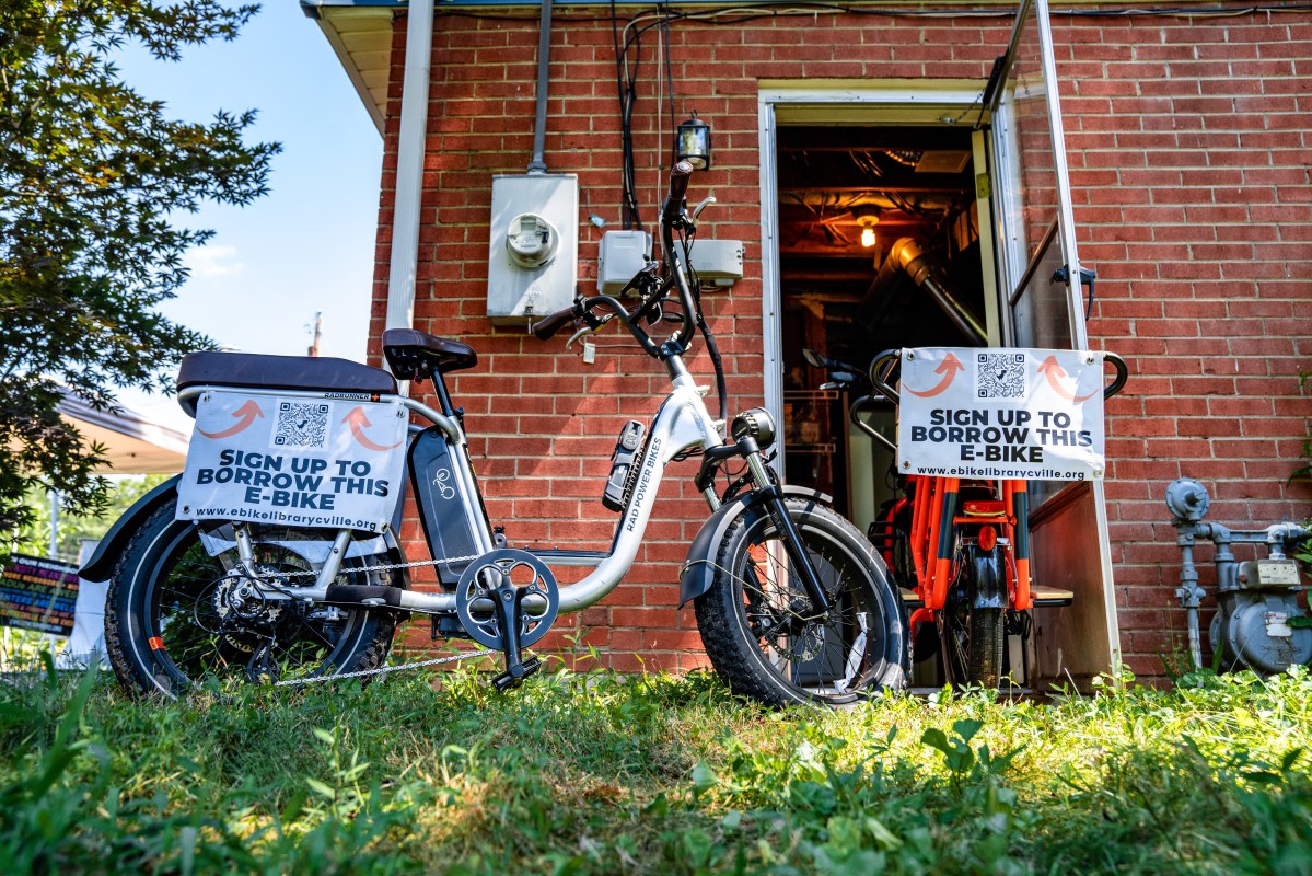 An e-bike is pictured in front of a brick wall with a door leading into a laundry room. Inside the door is a second e-bike. Both has signs attached that say, "Rent this e-bike."
