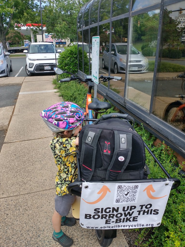 An e-bike is parked on the sidewalk beside a wall made entirely of windows. A sign attached to the back of the bike frame reads “sign up to borrow this e-bike, www.ebikelibrarycville.org.” A young child wearing a helmet stands next to the bike.
