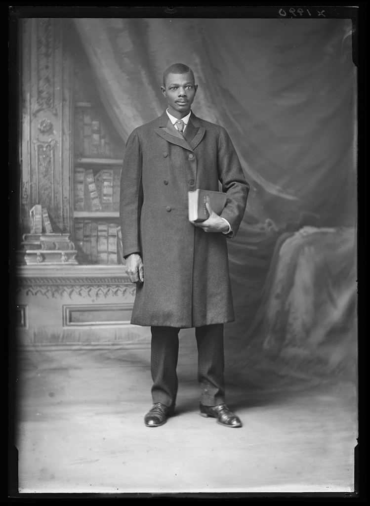 A black and white full-length portrait shows a man standing in front of a faux-library backdrop and looking directly at the camera. He wears a well-tailored long coat, his slacks and shoes visible underneath, and holds a thick book to his torso.