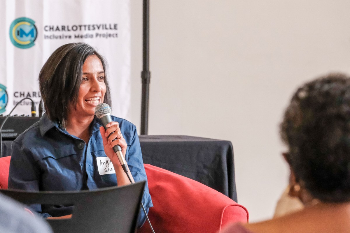 A woman sits on a red chair holding a microphone, with the head of an audience member in view. A sign behind her reads, “Charlottesville Inclusive Media.”