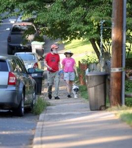 Two people with hats on walk a dog down a sidewalk, with lawns and cars on either side.