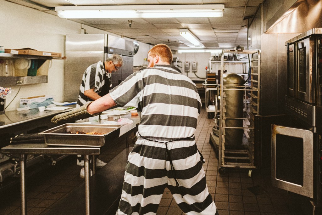 A man in a black and white stripped prison jumpsuit pulls a pan of food from an oven in an industrial kitchen full of stainless steal appliances.