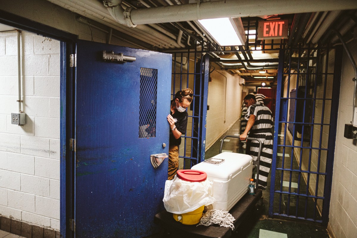 Men in black and white stripped prison jumpsuits wheel a large box down a prison hallway.