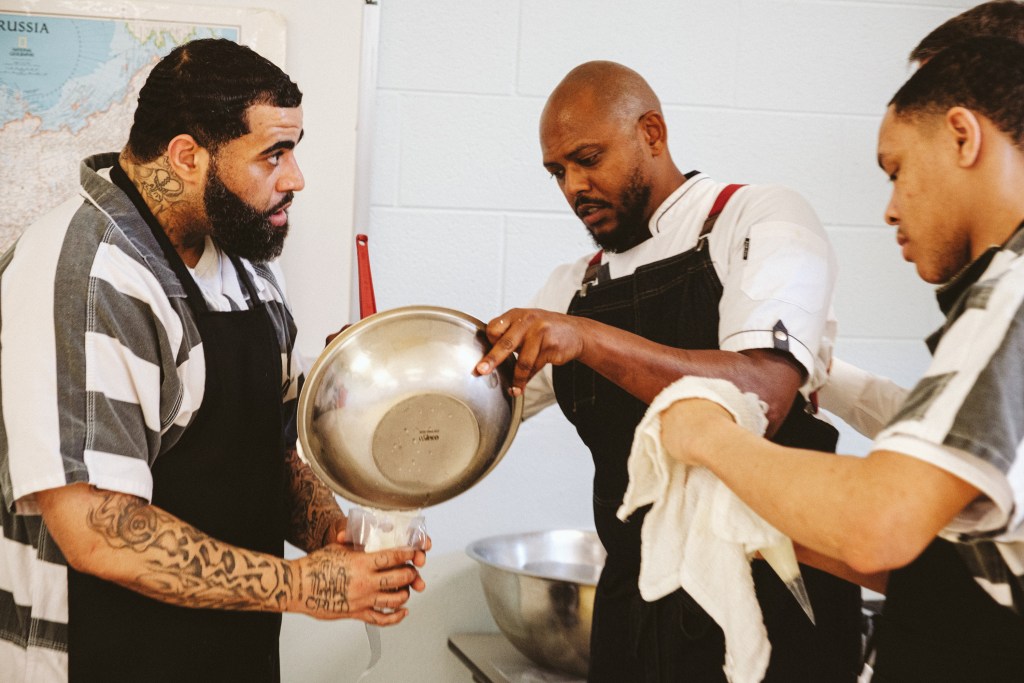At the center of the photo a man in a white chef's coat stirs something in a stainless steal bowl. On either side of him, two men in black aprons over black and white striped jumpsuits watch what he is doing.