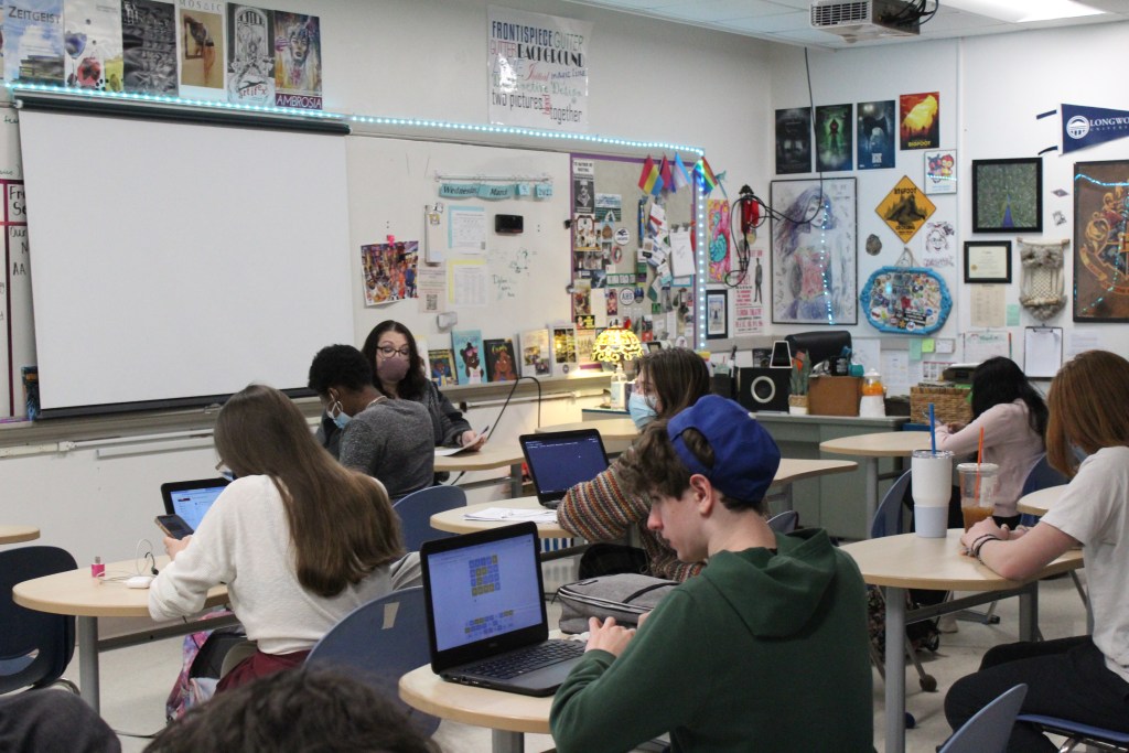 Teenagers sit at desks in a classroom facing a whiteboard. An adult sits at a desk facing them.