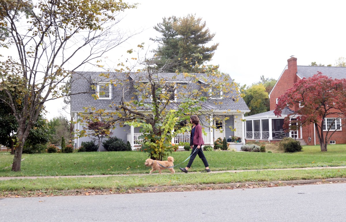 A woman walks a dog down a sidewalk in front of a while house with a large yard.