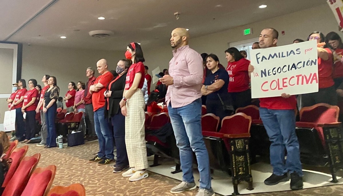A group of people are pictured standing in an auditorium. Some hold signs, many are wearing red shirts.