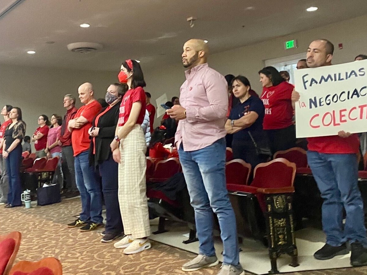 A group of people are pictured standing in an auditorium. Some hold signs, many are wearing red shirts.