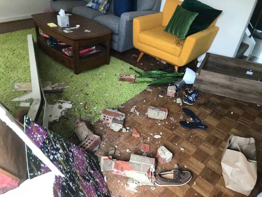 A living room with a yellow chair, gray sofa, coffee table and green rug is covered in debris and bricks, with household items strewn around.