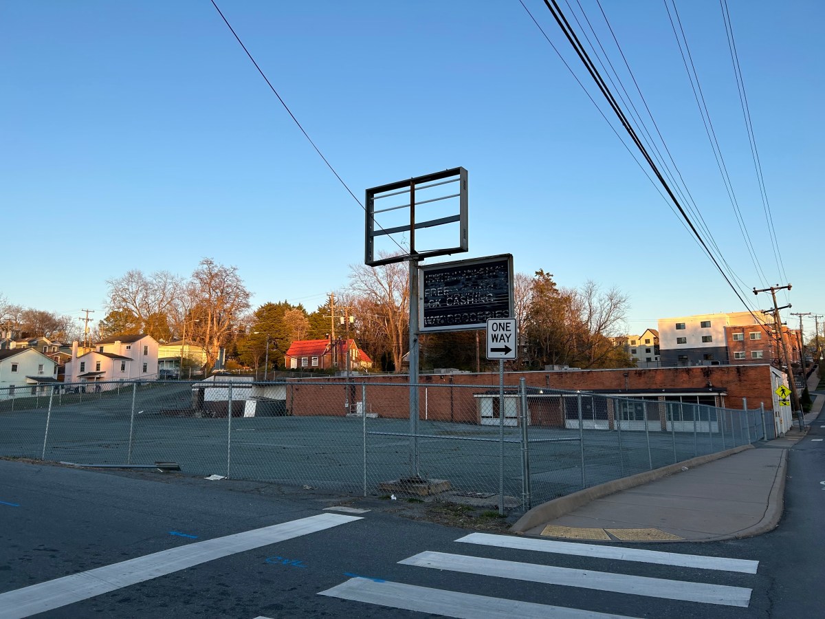 A lot surrounded by a wire fence at the corners of a street, with a one-story brick building. A sign in front is empty, except for some washed out text.