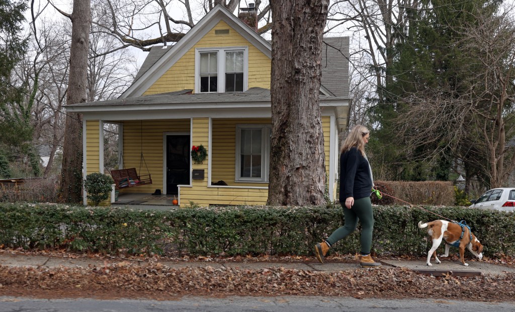 A woman walks a dog on a sidewalk in front of a yellow house, with fallen leaves  lining the sidewalk.