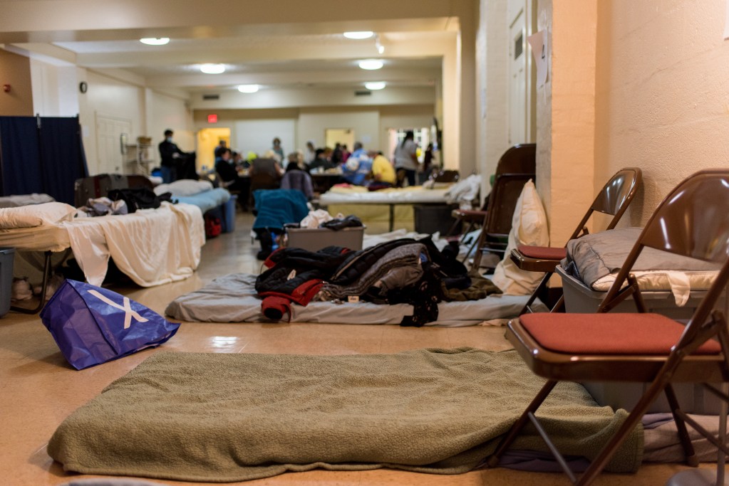 A room with linoleum floors and fluorescent lights is filled with cots and bed rolls.