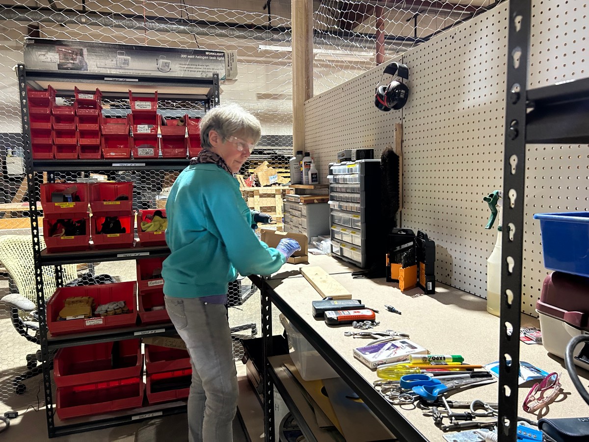 Woman in teal sweater stands at work bench with tools in front of her, shelves of organized parts to the side.