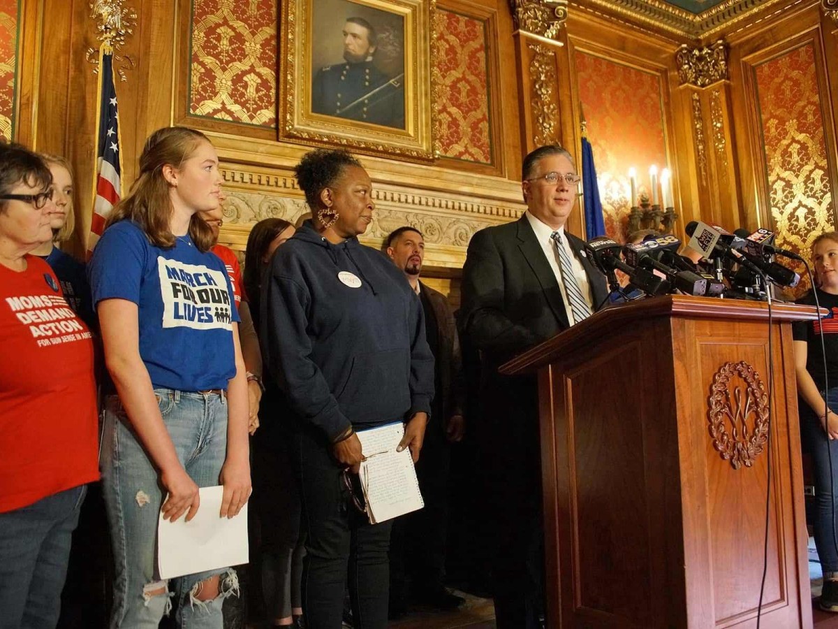 People lined up next to a podium with a man in a suit speaking at microphones, two on the edge are wearing red and black t-shirts with slogans on them. There is a historical painting behind them.