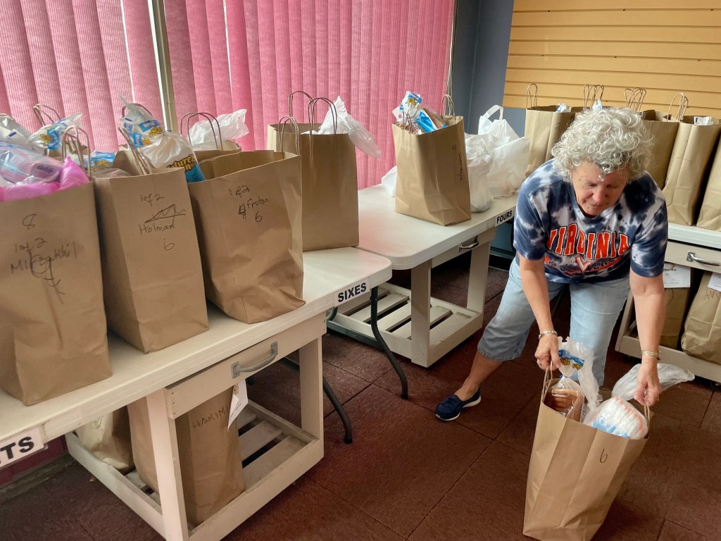 A woman lifts a paper bag of groceries onto a table filled with dozens of identical bags.