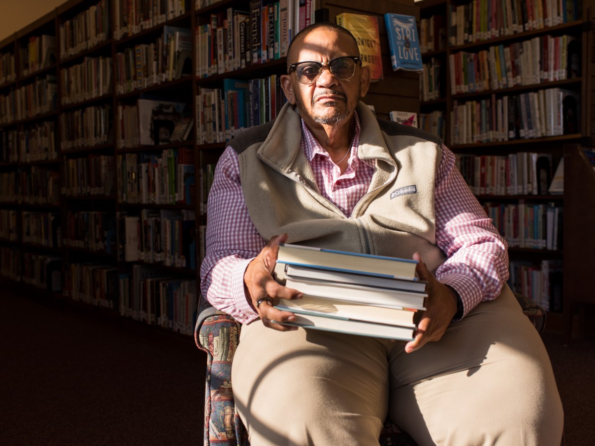 A person in a chair holding a stack of books, in front of shelves of books, with a band of light across his face.