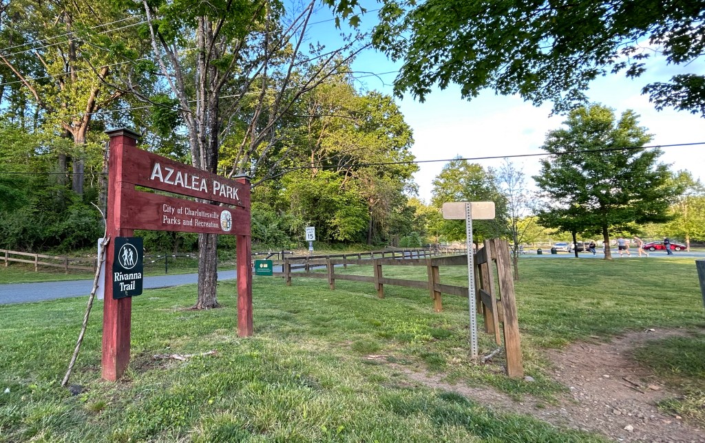 A wooden sign reading "Azalea Park: City of Charlottesville Parks and Recreation" is the focus of the photo. A smaller sign attached to it reads "Rivanna Trail." A branch, possibly used as a walking stick, leans up against one side of the wooden sign. Nearby there is a wooden fence and grass with a dirt trail, and in the distance, a few people play a pickup basketball game in a court near a parking lot. The trees are full and the grass is lush.